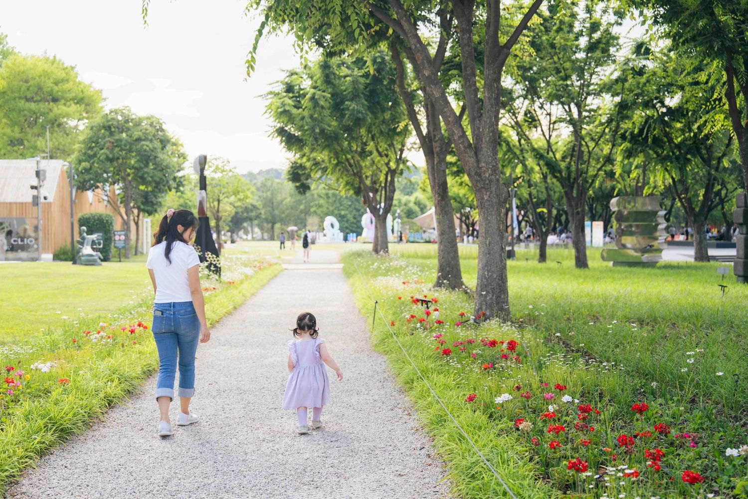 A mother and young daughter walk together along a scenic park path lined with colorful flowers and lush green trees.