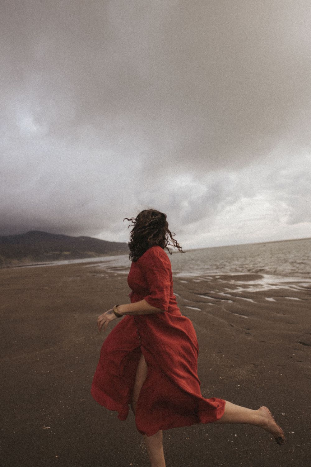 A woman in a flowing red dress runs barefoot along a stormy beach with dramatic cloudy skies and ocean waves.