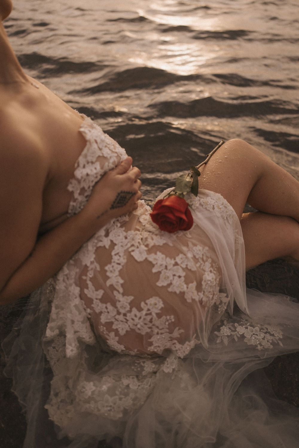 Pregnant woman in white lace gown holding a red rose, sitting by ocean waves at golden hour.