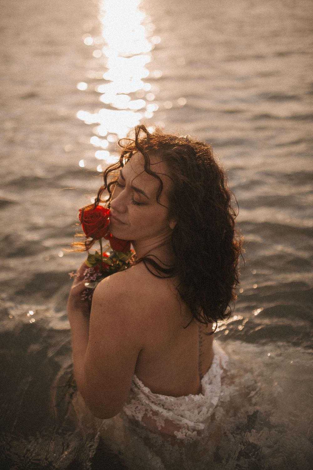 Woman in white lace dress holding a red rose while standing in shimmering golden water at sunset.