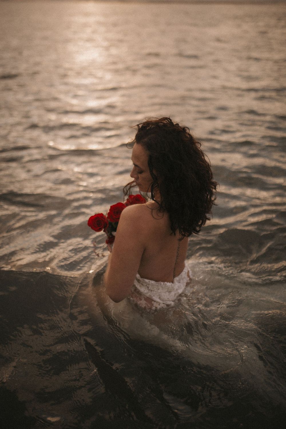 Woman in white dress holding red roses standing in ocean water at golden sunset.