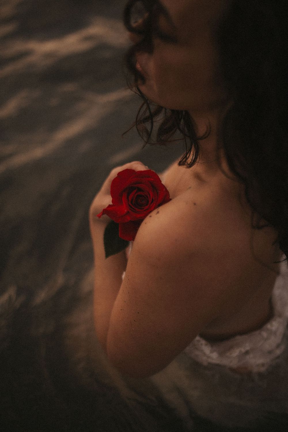 A woman holds a red rose against her bare shoulder while partially submerged in dark, rippling water.
