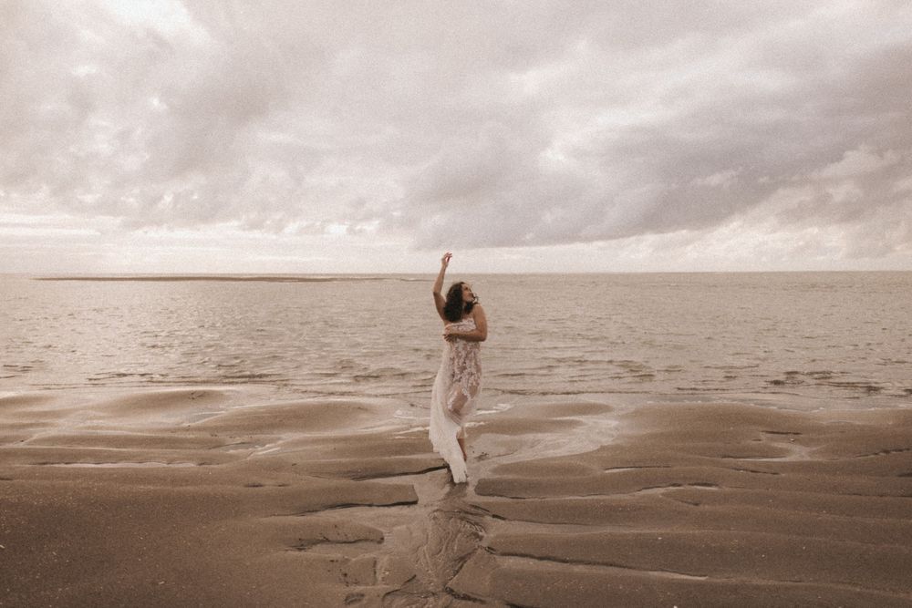 A woman stands on a sandy beach with arms raised, facing a calm sea under a dramatic cloudy sky.