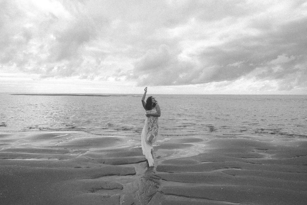 Black and white photo of a woman in a dress standing on a sandy beach with arms raised under a cloudy sky.
