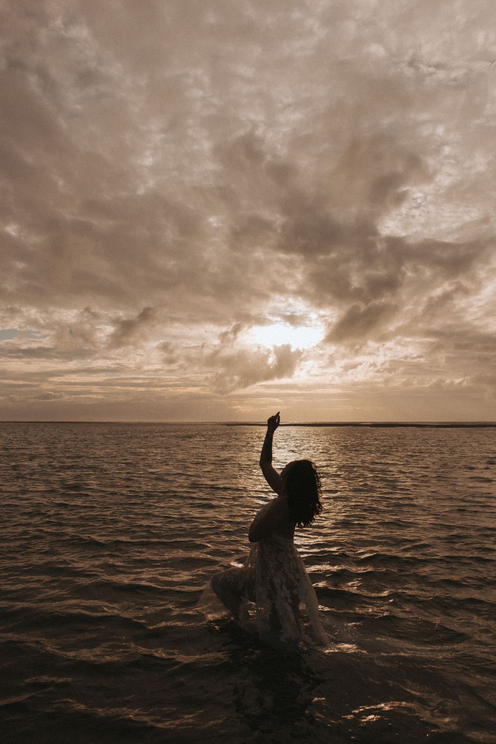 Silhouette of a woman in a dress standing in ocean water with arm raised against a dramatic sunset sky.