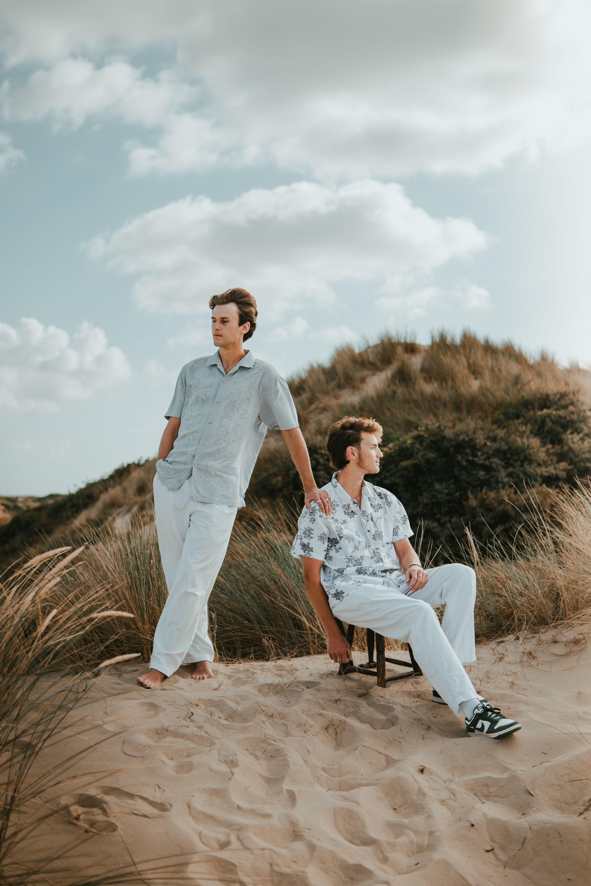 Two young men pose on sandy beach dunes, one standing and one seated, wearing casual summer outfits under a cloudy sky.
