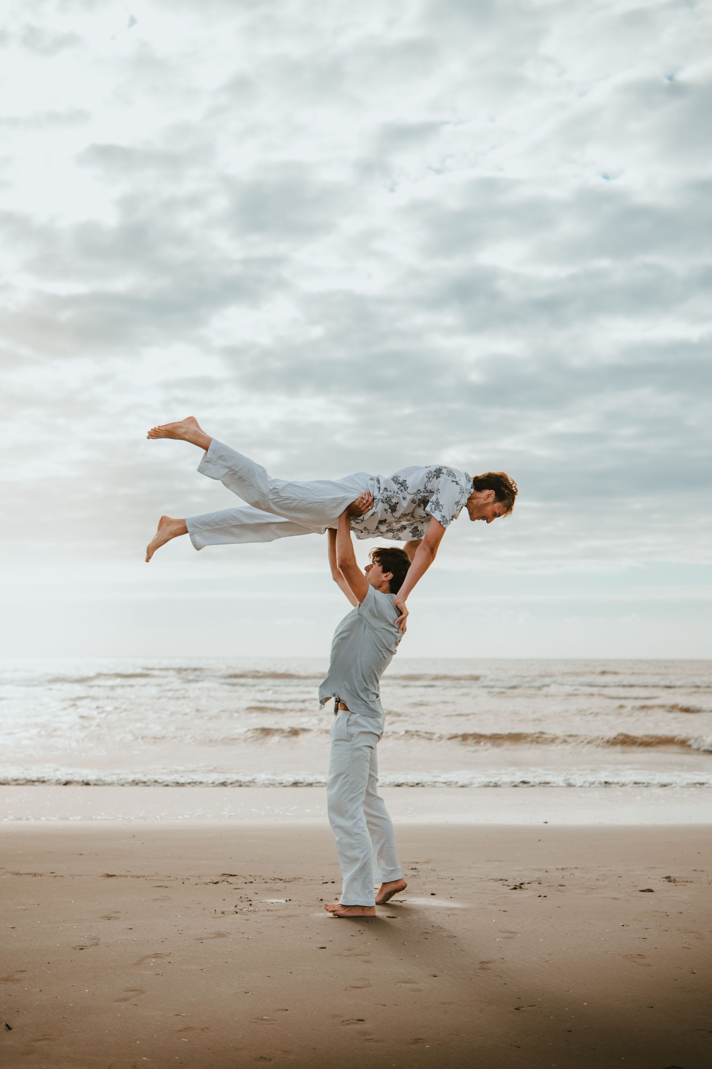 Couple performing acrobatic lift on sandy beach with ocean waves and cloudy sky backdrop.