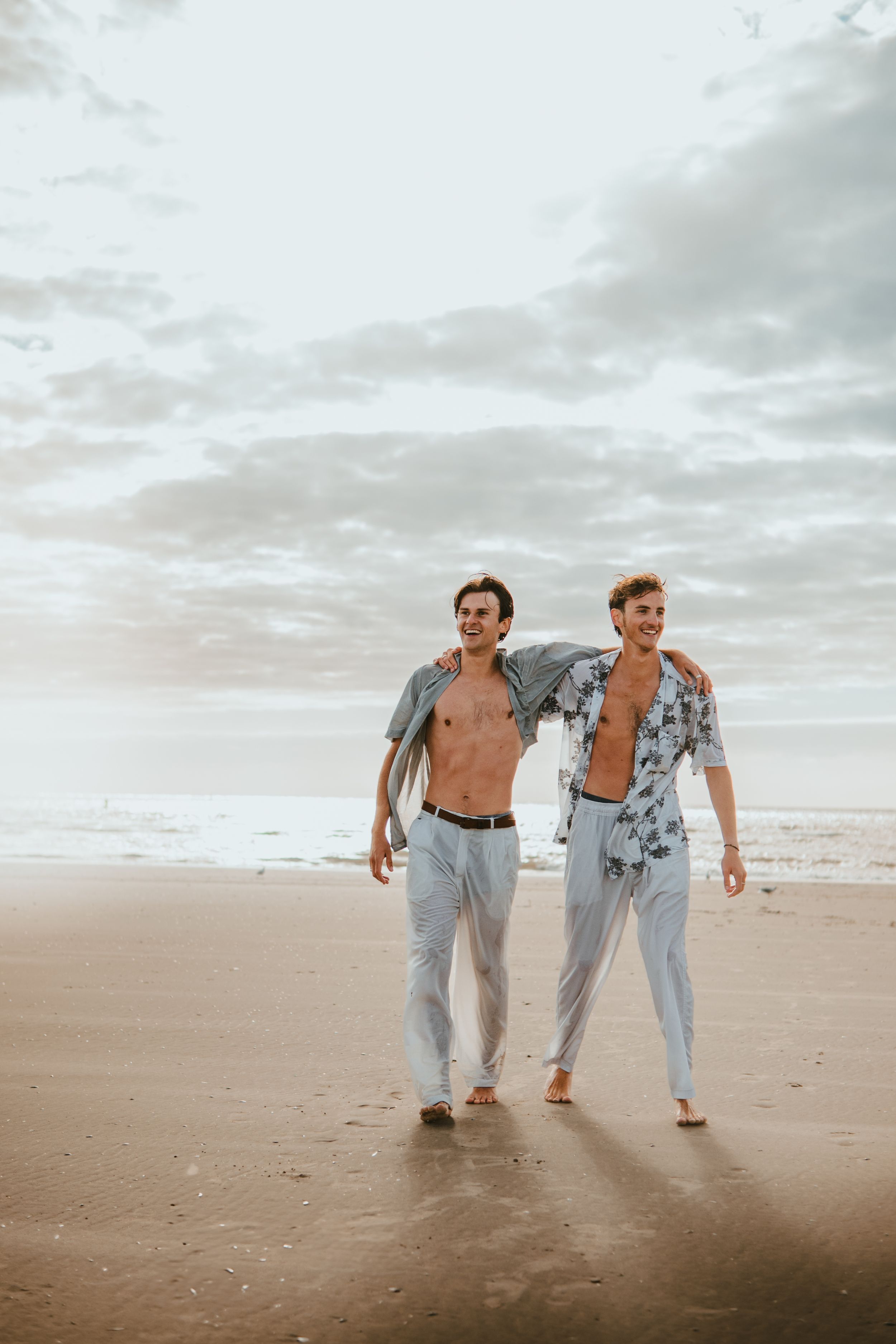 Two young men walking barefoot on a sandy beach with open shirts and light pants under a cloudy sky.