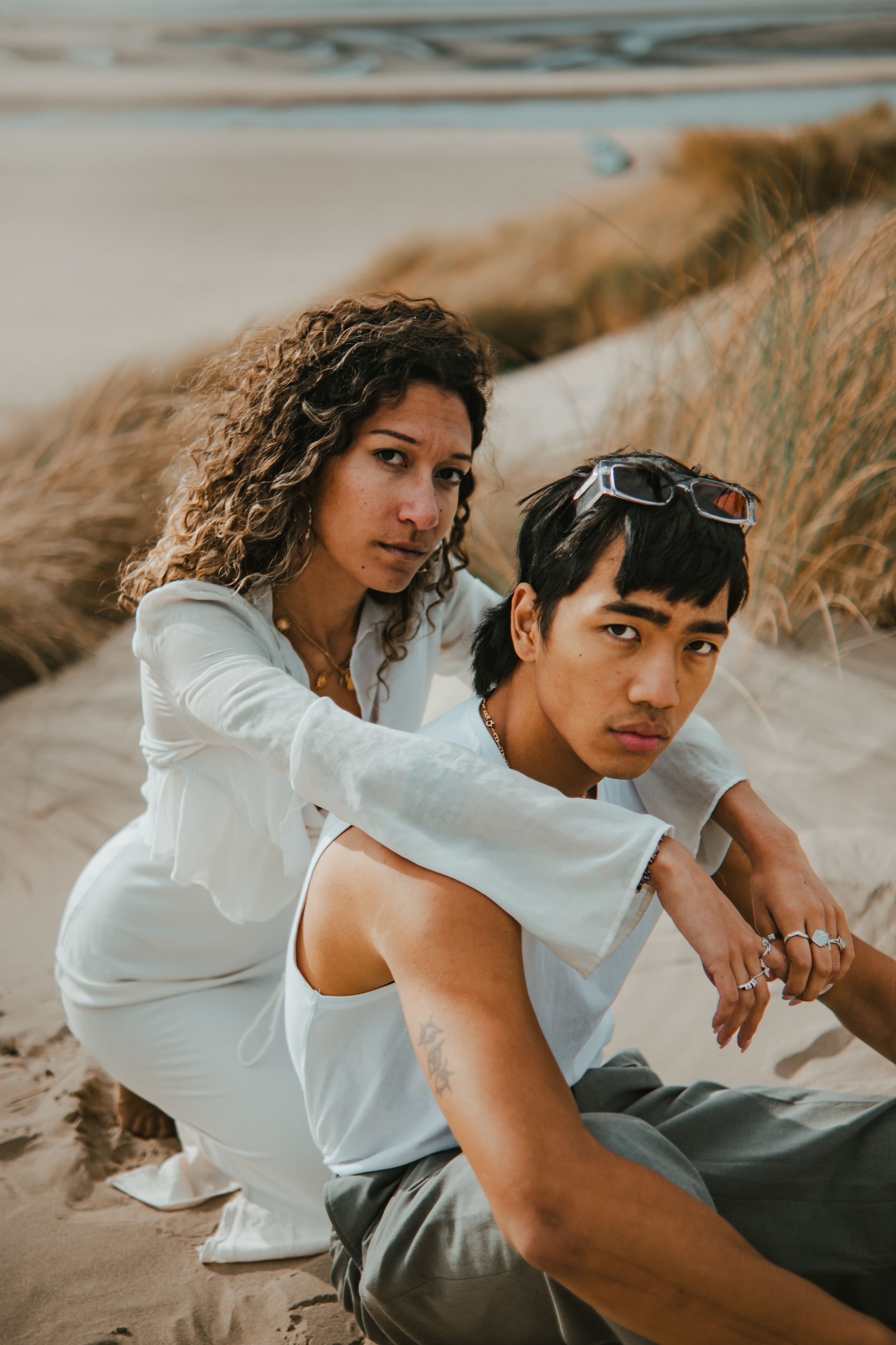 Two young people in white outfits sitting on sandy beach dunes, woman with curly hair and man with sunglasses on head.