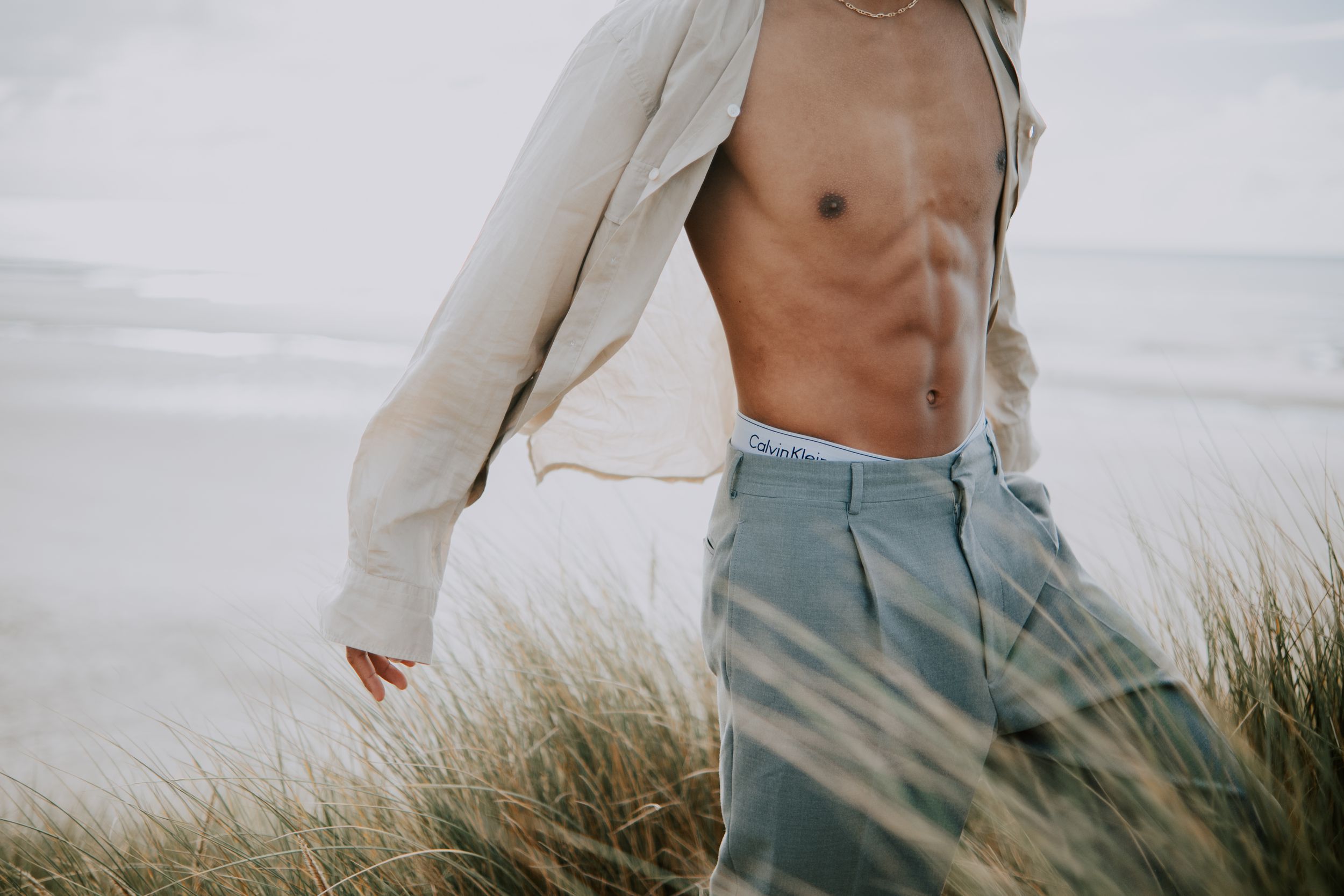 Shirtless man with toned abs in open linen shirt and blue jeans standing in beach dune grass.