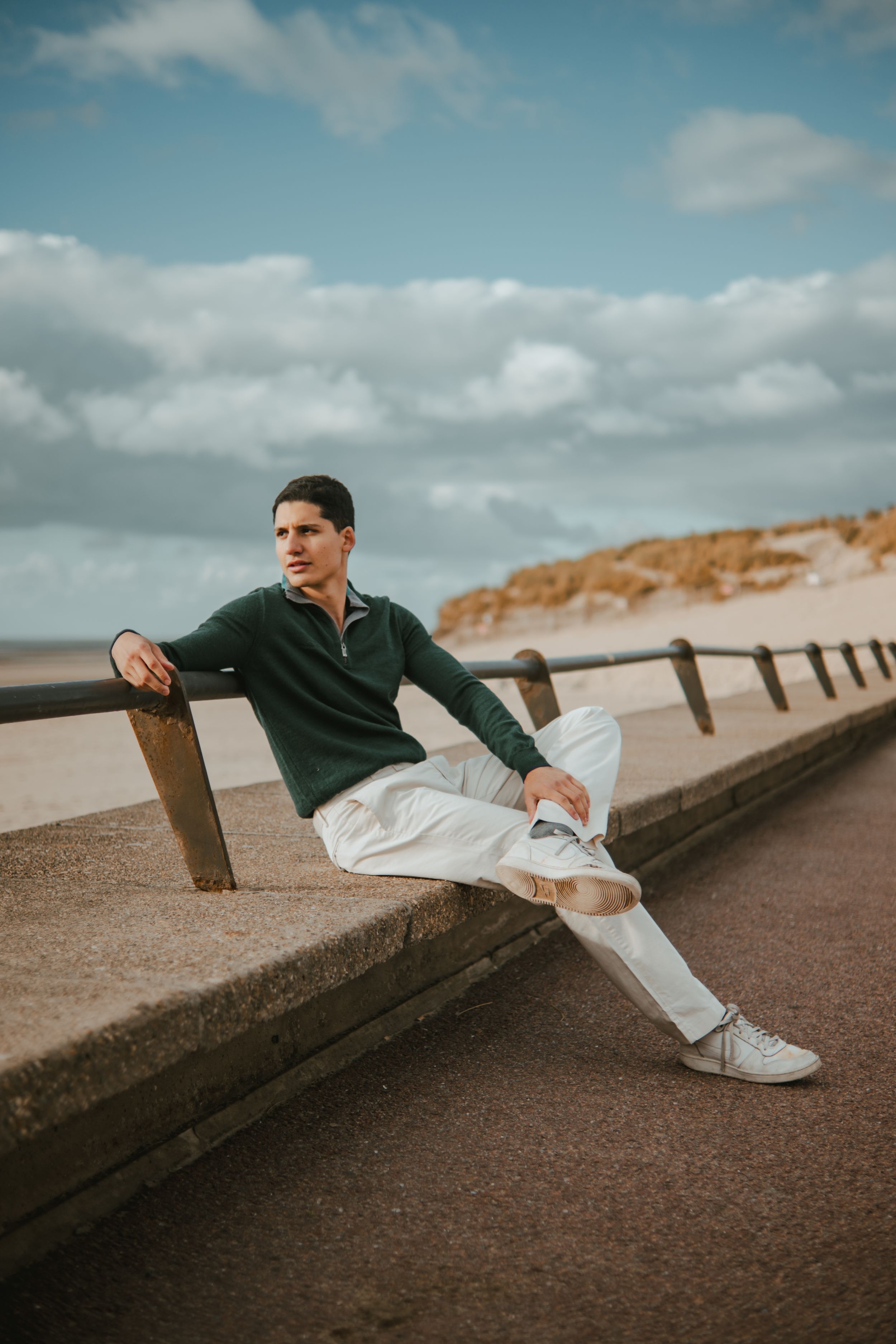 Young man in green sweater and white pants sitting on a seaside promenade railing with sandy dunes behind him.