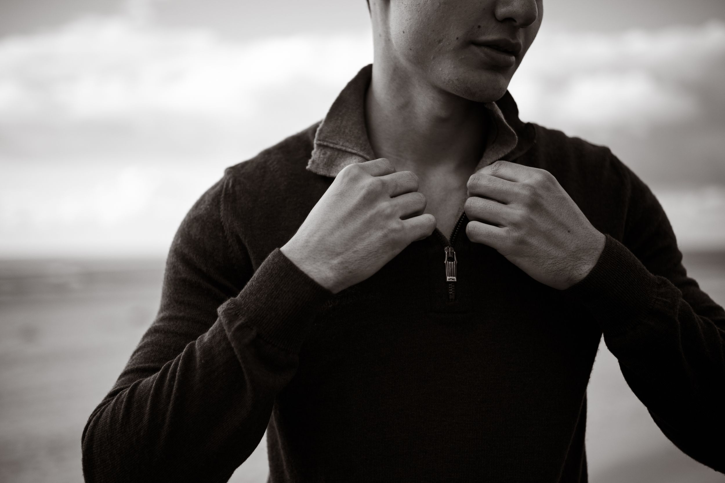 Black and white portrait of a young man adjusting the collar of his zip-up sweater outdoors.