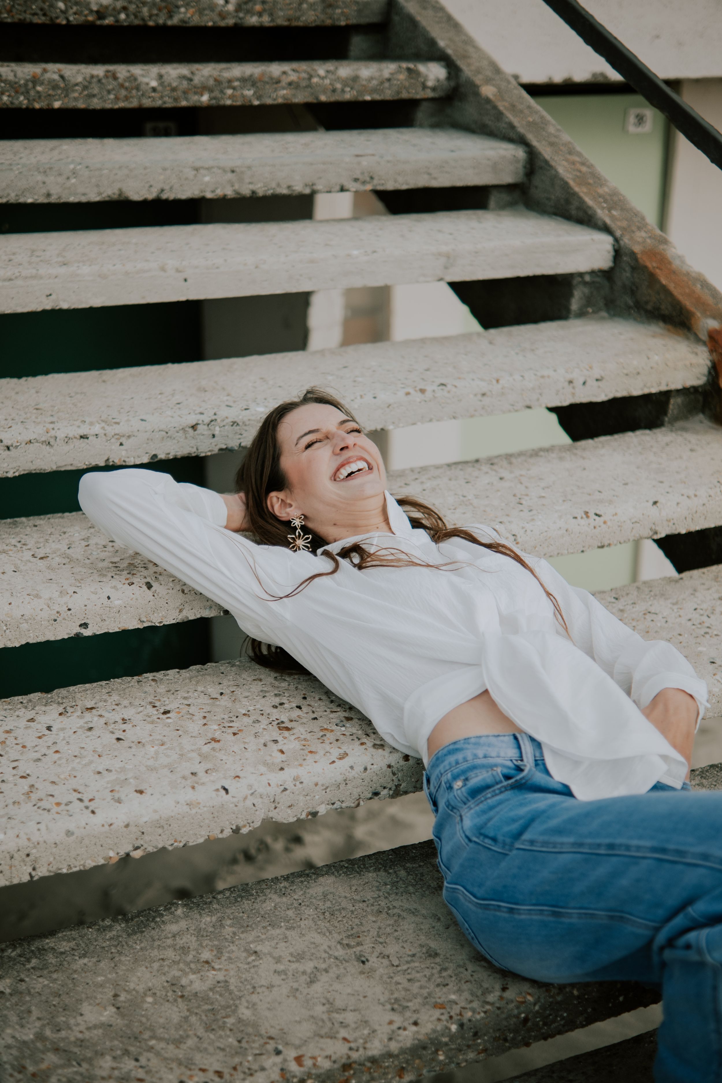 Young woman in white shirt and jeans laughing while lying on concrete outdoor steps.