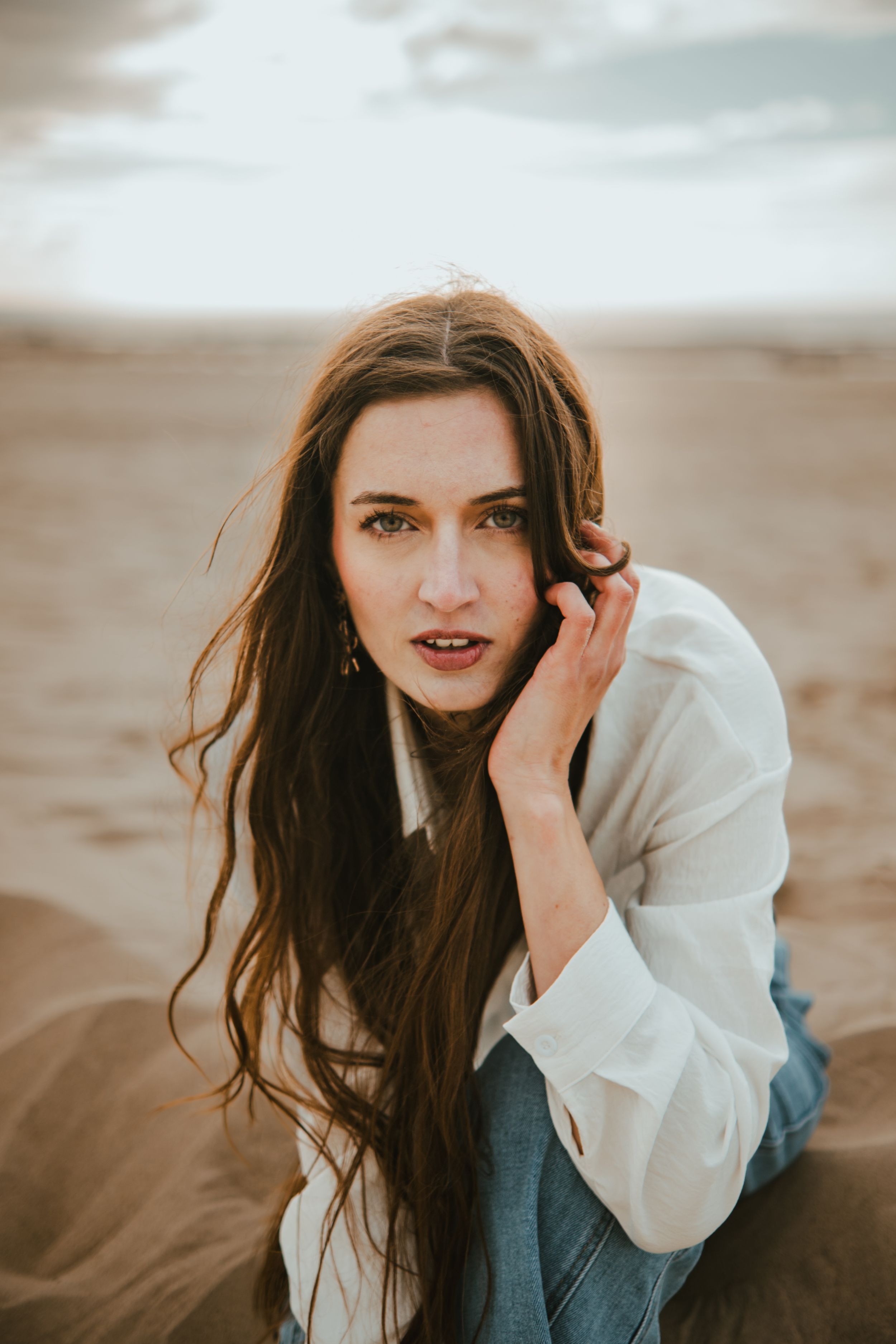 Young woman with long brown hair sitting on sandy beach, wearing white shirt and jeans, gazing at camera.