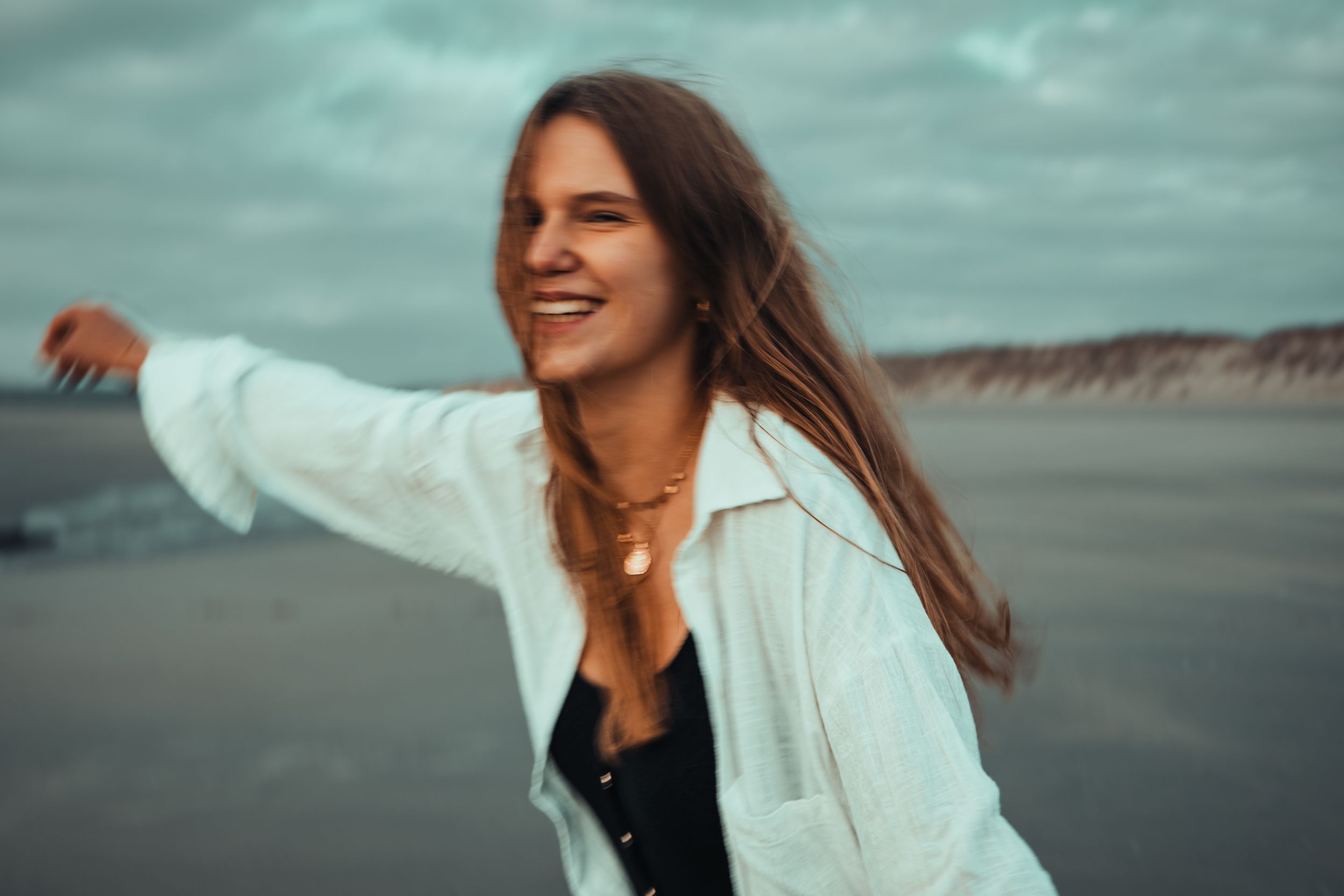 Smiling young woman with long hair wearing a white jacket and gold necklace on a beach with cloudy skies.