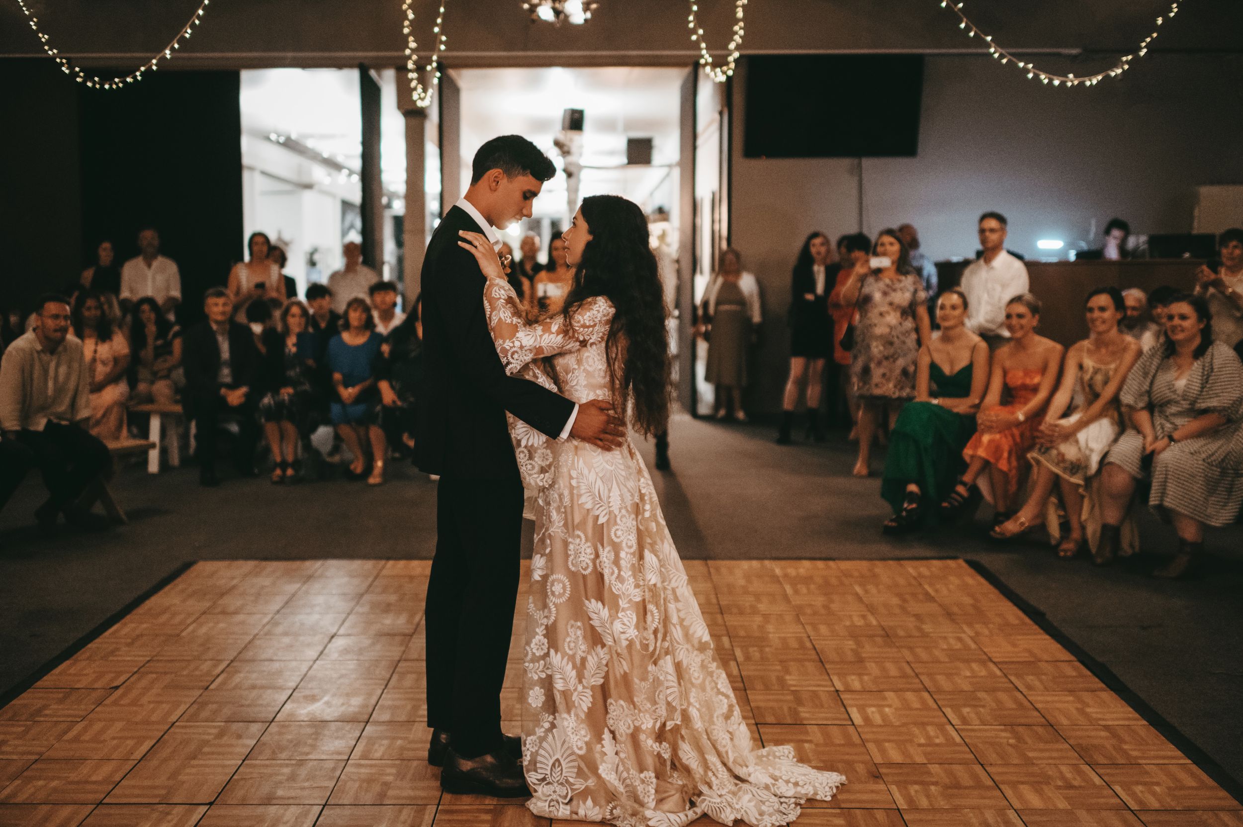 Bride and groom share first dance at wedding reception with guests watching in dimly lit venue with string lights.