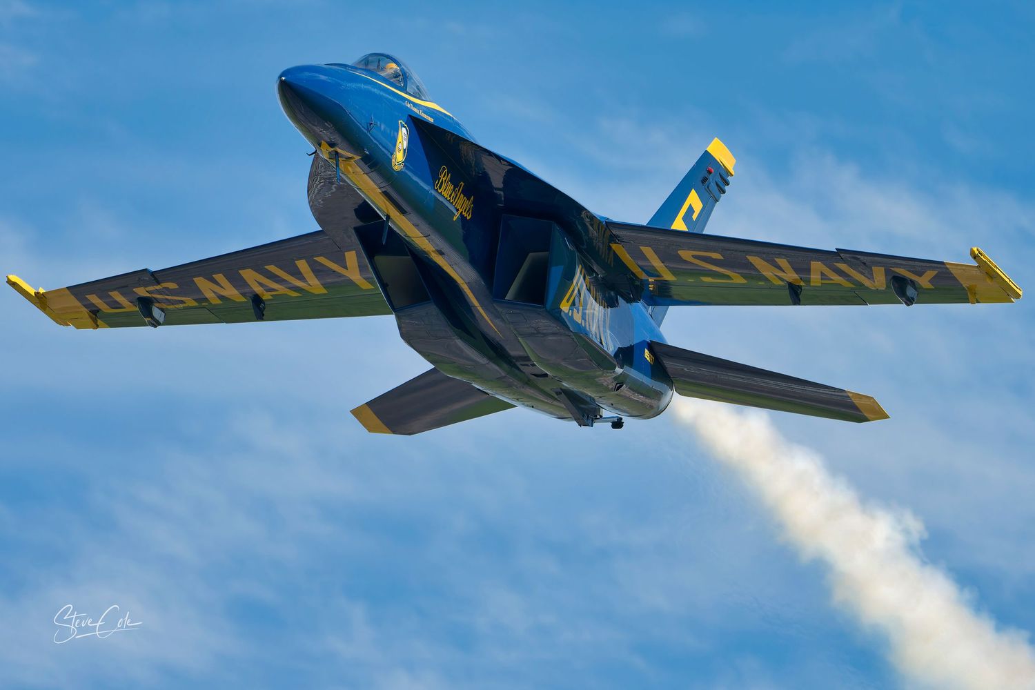 Blue Angels F/A-18 jet in a steep climb displaying "US NAVY" lettering under the wings, photo by Steve Cole Photography.