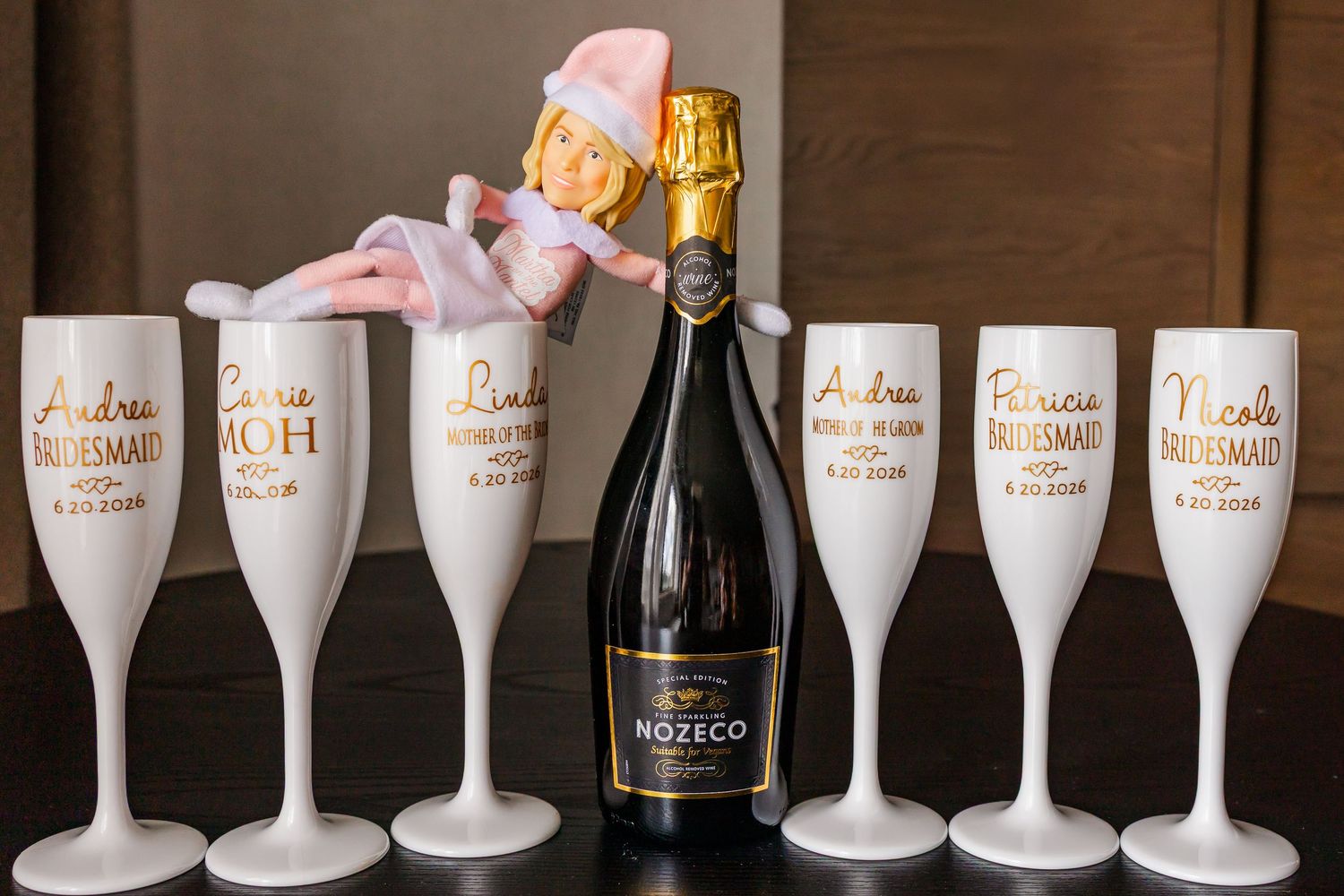 Personalized white champagne flutes with gold text surround a Nozeco bottle, with a decorative pink ballerina figurine.