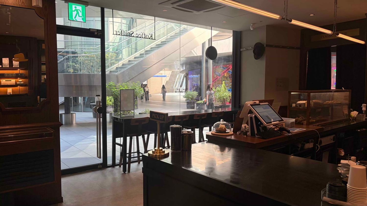 Modern cafe interior with dark wood bar, glass facade, and lush outdoor greenery visible through floor-to-ceiling windows.