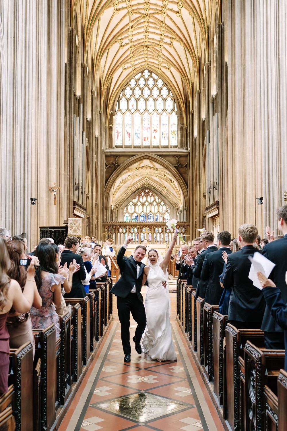 Joyful newlyweds walk down the aisle of a grand Gothic cathedral, celebrated by guests in elegant attire at St Mary Redcliffe Church Bristol