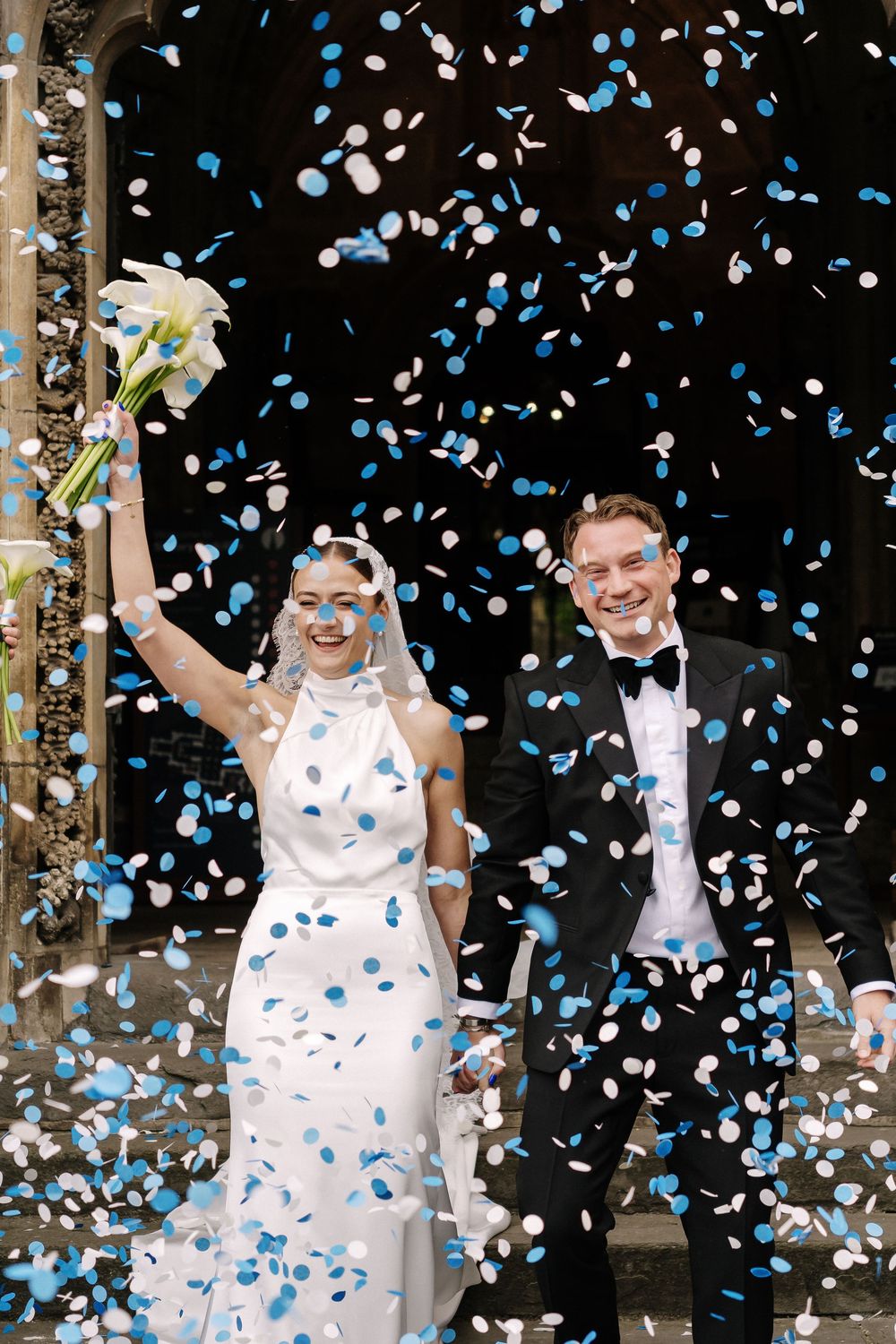 Joyful newlyweds celebrate outside church surrounded by blue and white confetti, bride in white gown holding bouquet aloft St Mary Redcliffe
