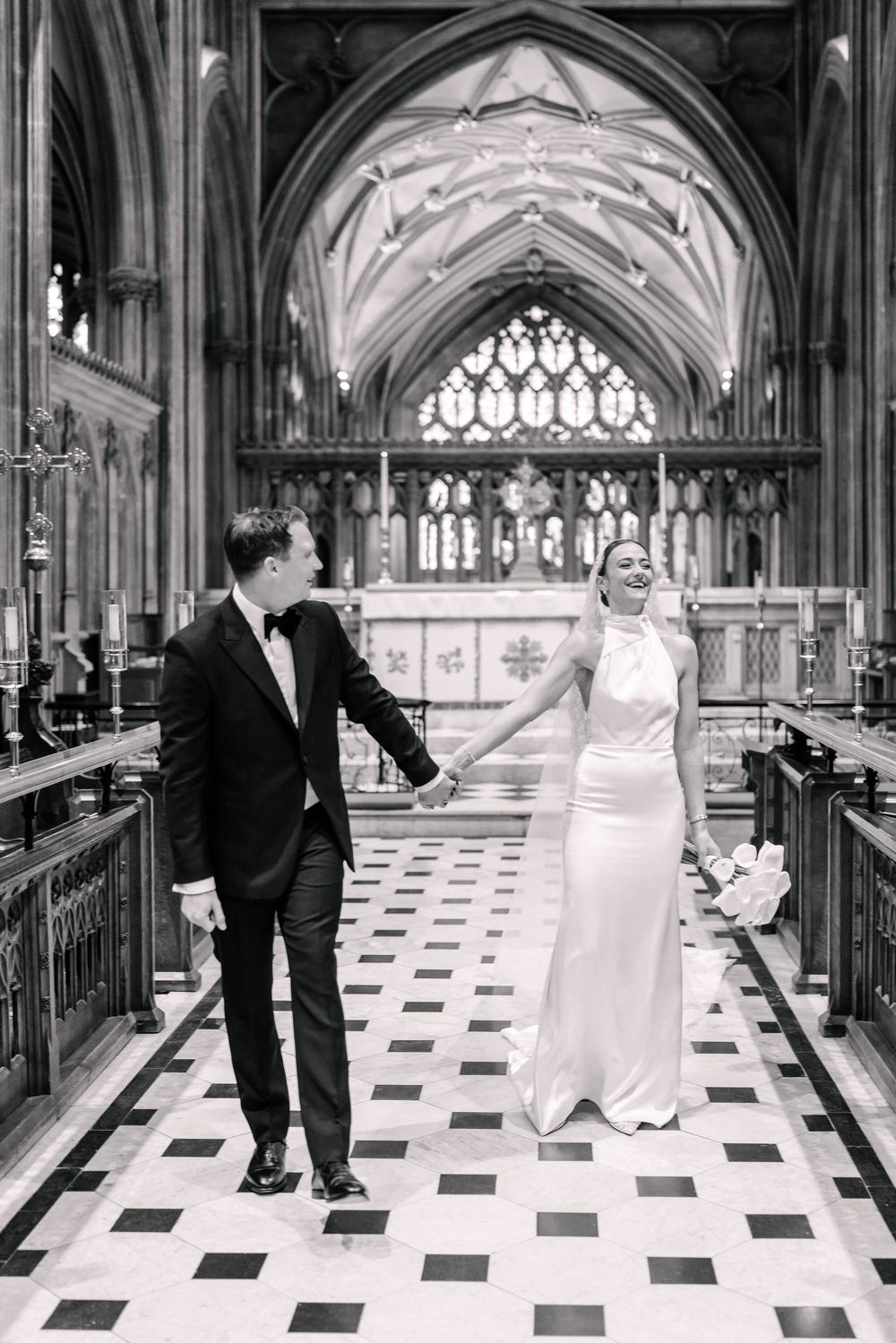 Black and white photo of a bride and groom holding hands walking down a the grand St Mary Redcliffe Church