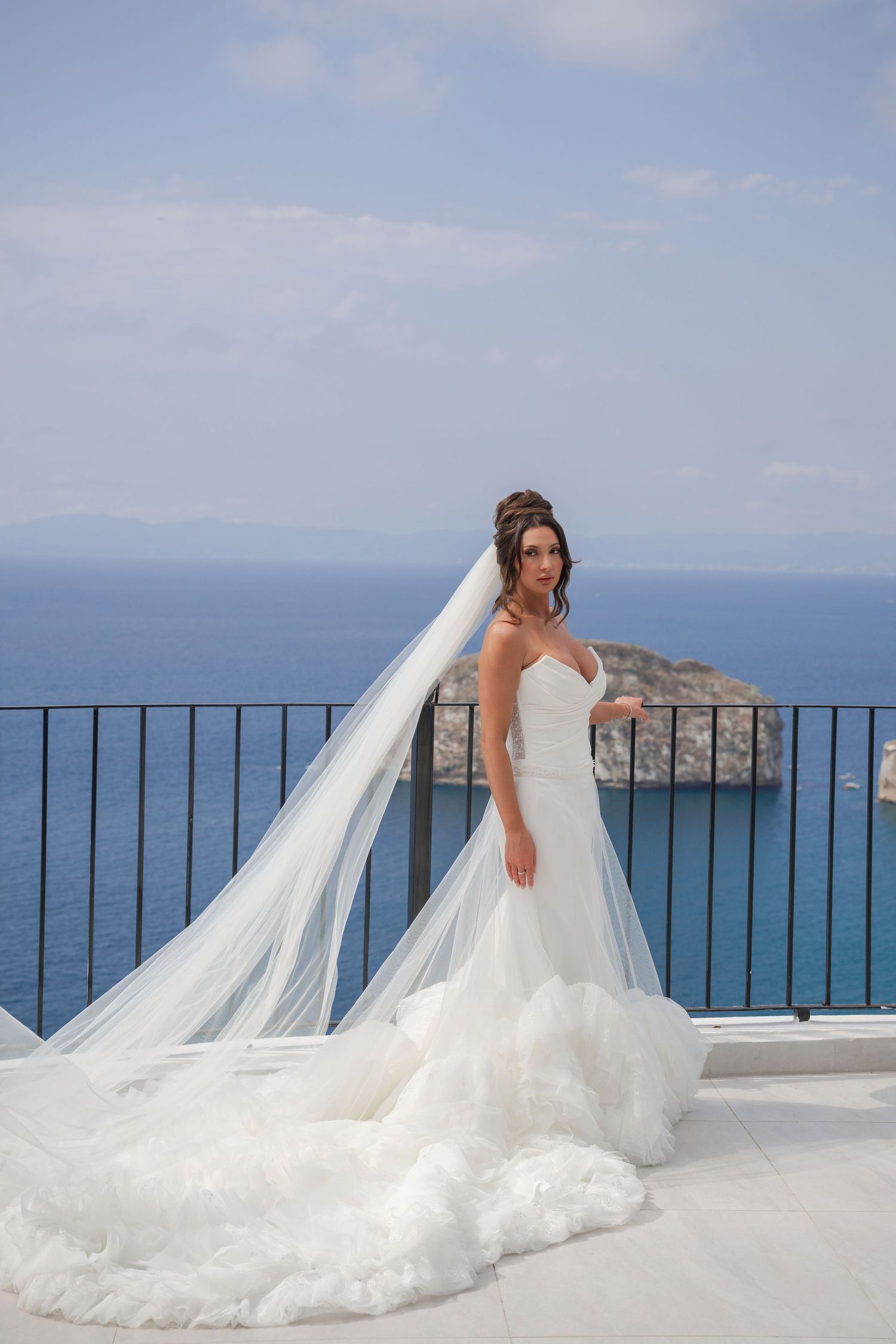 Bride in a strapless mermaid wedding gown with a flowing cathedral veil poses on a terrace overlooking the blue sea.
