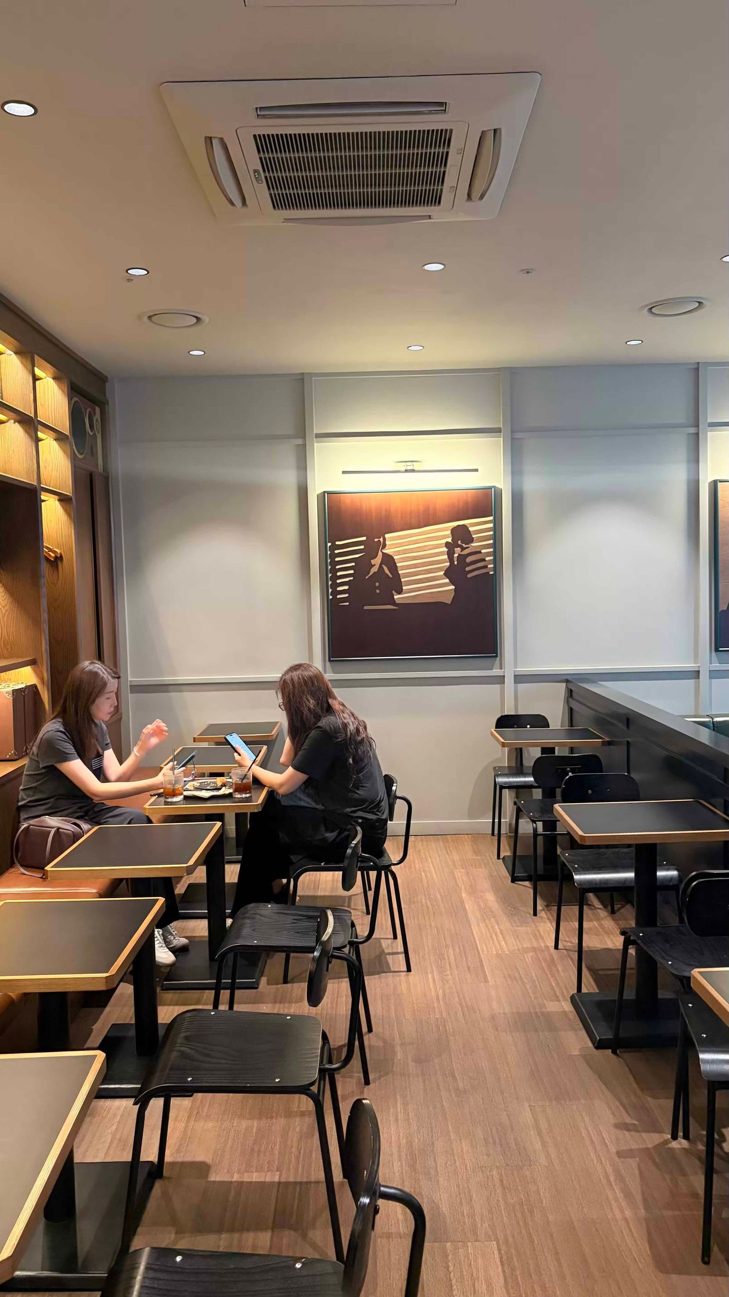 Two women sitting at a table in a modern restaurant with wooden floors, black chairs, and artwork on the wall.
