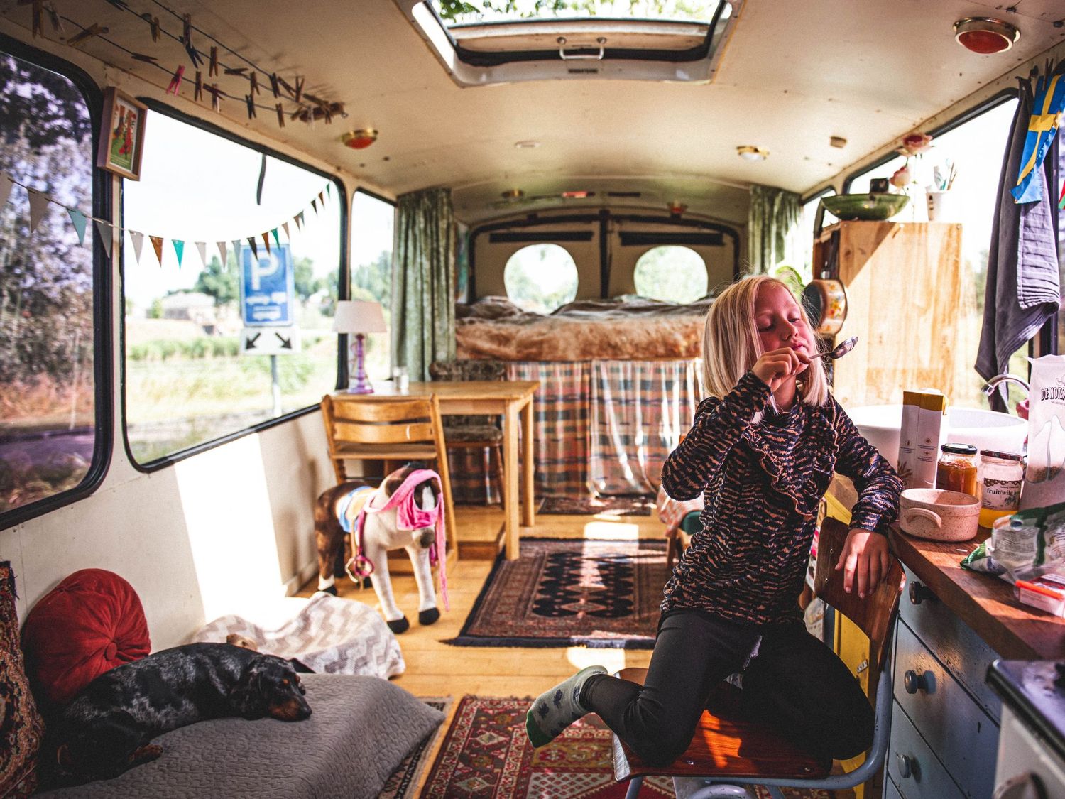 Woman sitting inside a converted bus home with colorful rugs, bunting, and cozy interior decor.