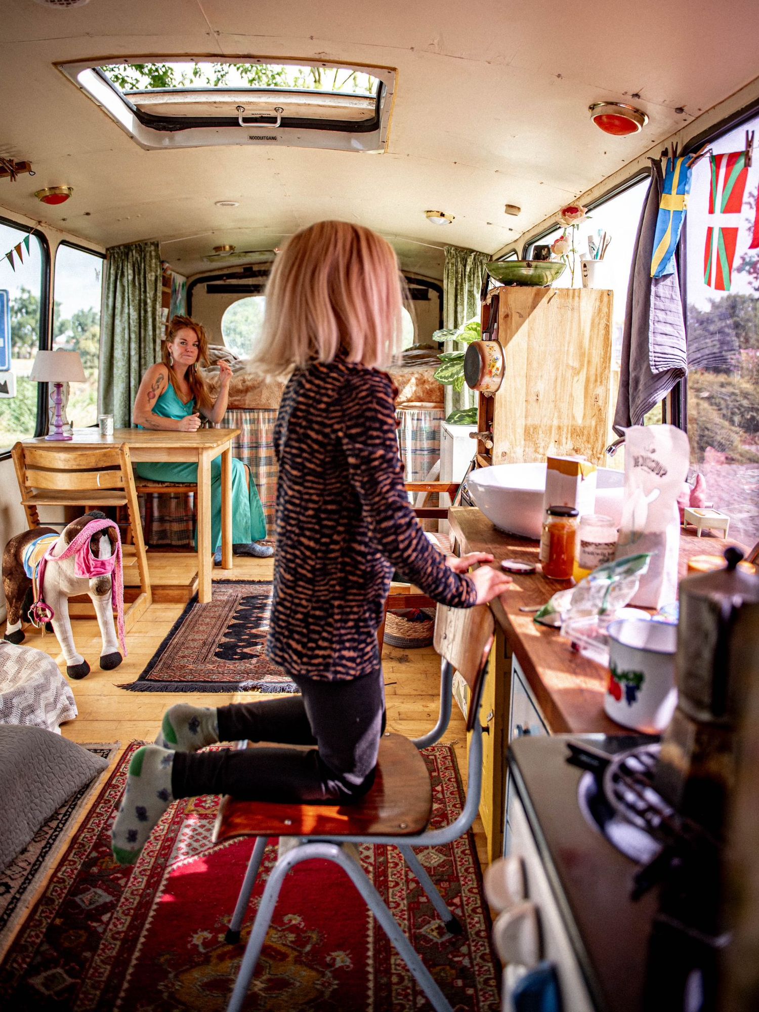 Child kneeling on stool at counter inside a converted school bus home with colorful rugs and flags.