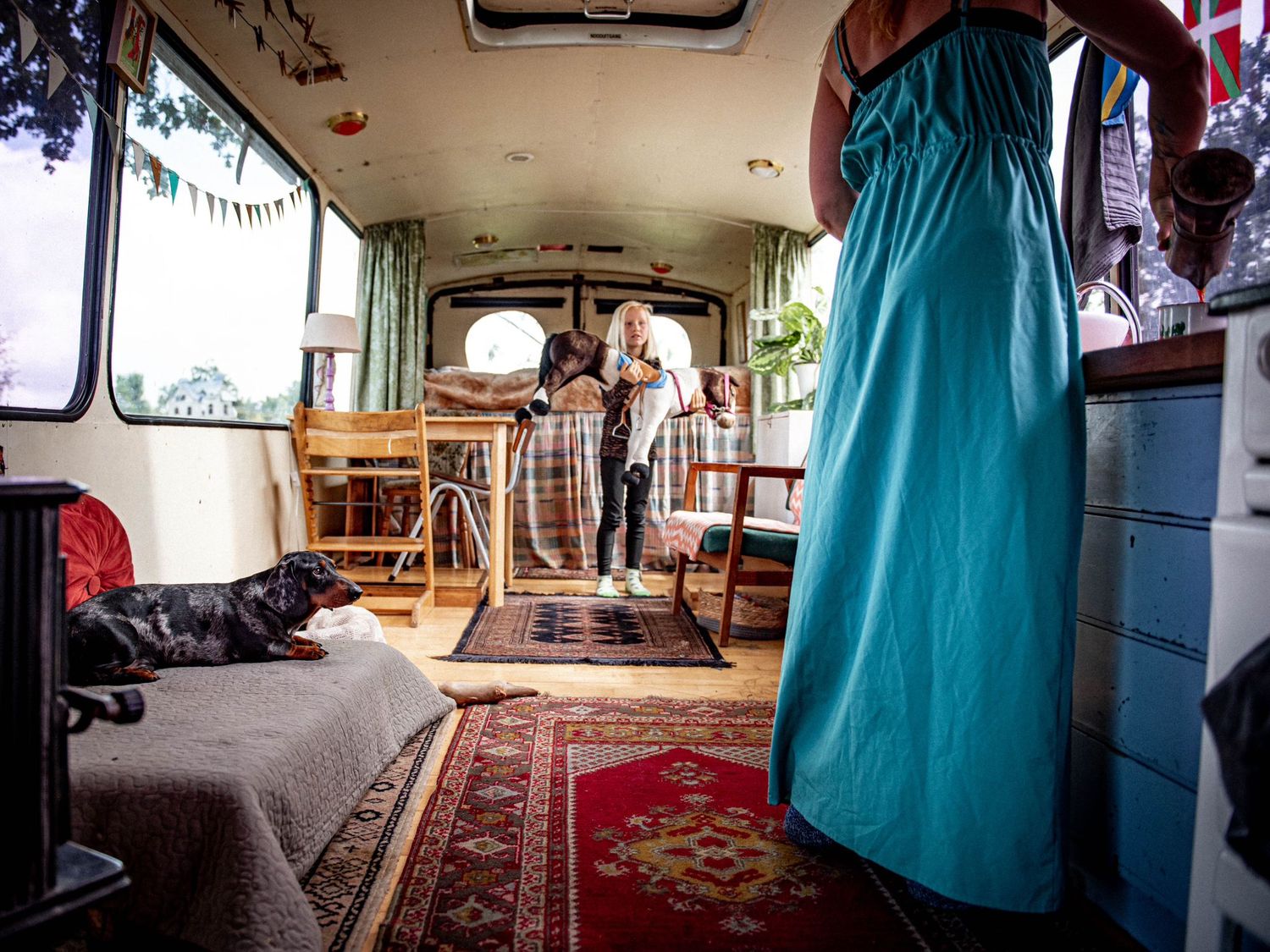 Interior of a converted bus home with a dog resting on a bed, colorful rugs, and a person in a teal dress.