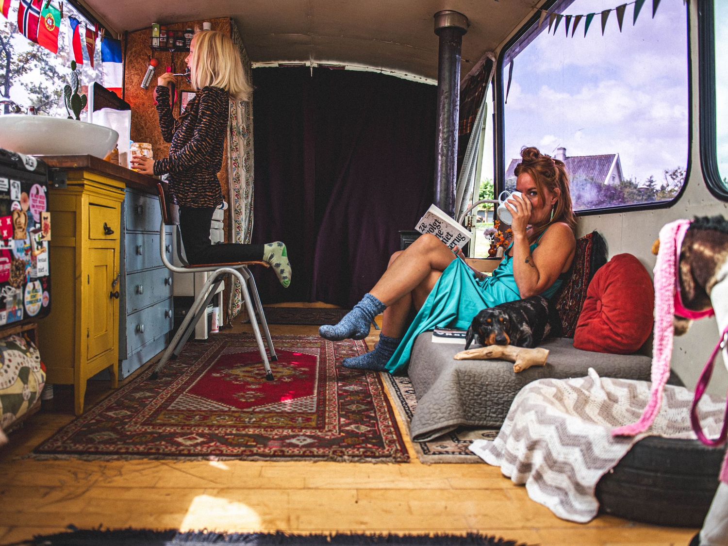 Two women relaxing inside a colorfully decorated vintage camper van with rugs, cushions, and eclectic decor.