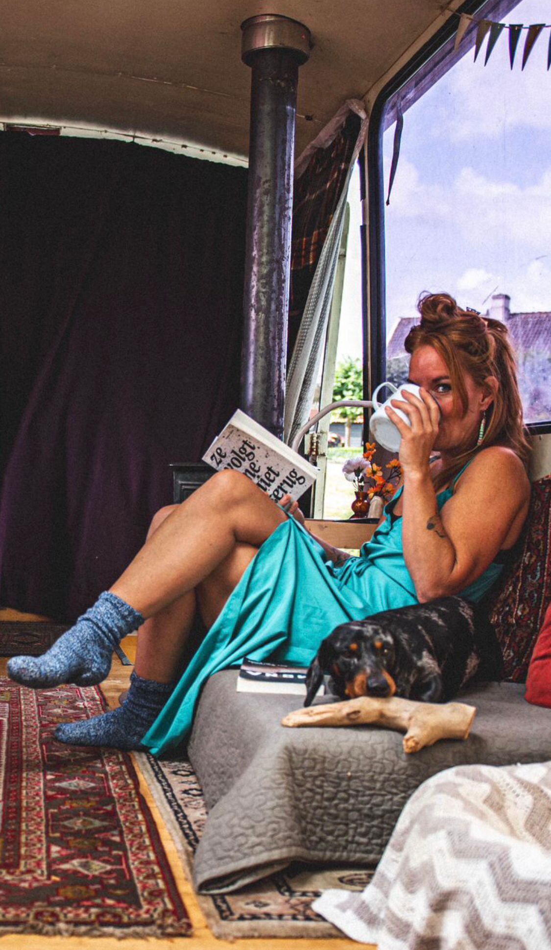 Woman in teal dress and blue socks relaxing with coffee and book beside a dachshund dog inside a cozy converted bus.