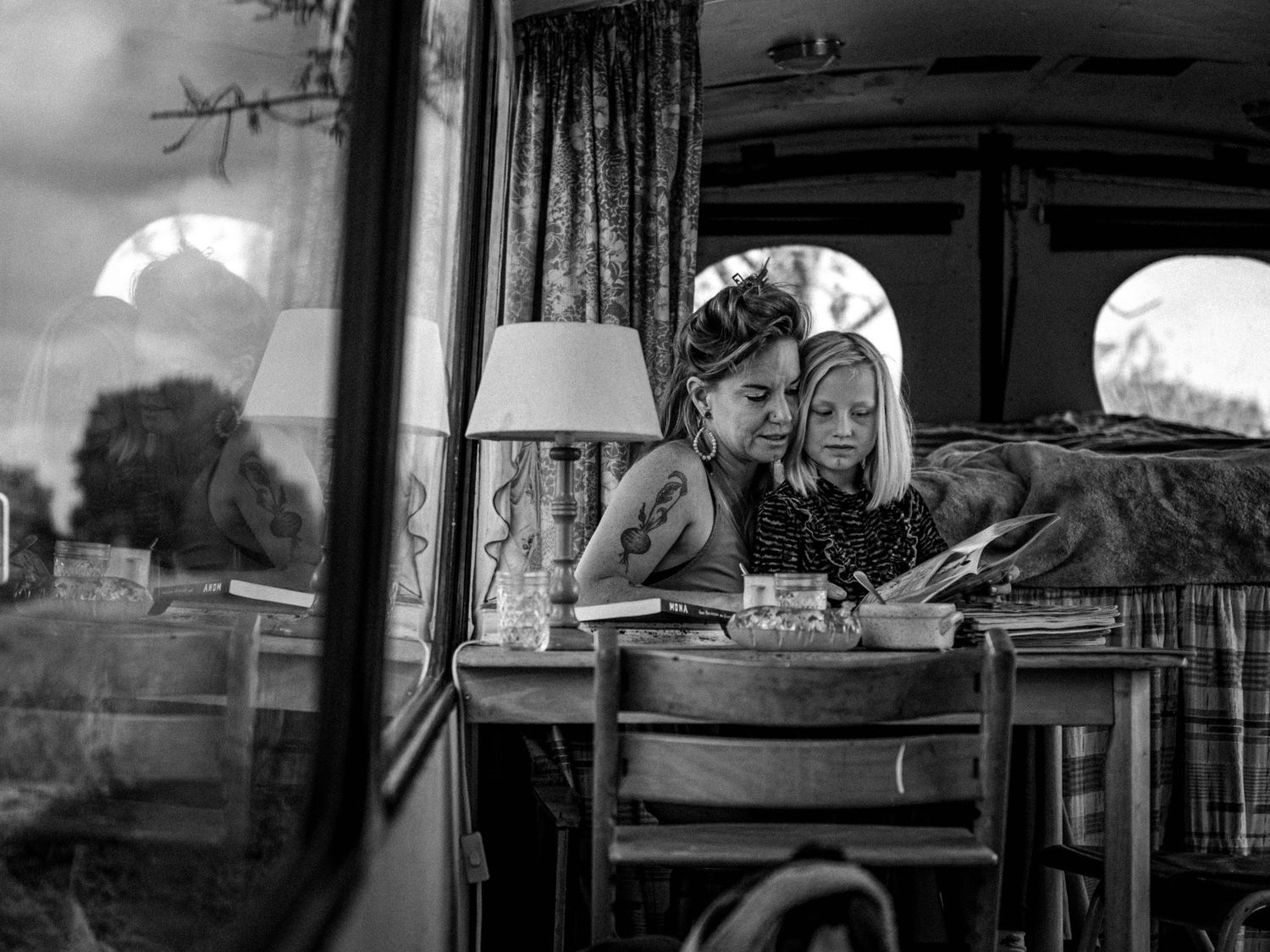 Black and white photo of two children reading together at a wooden table inside a vintage converted bus interior.