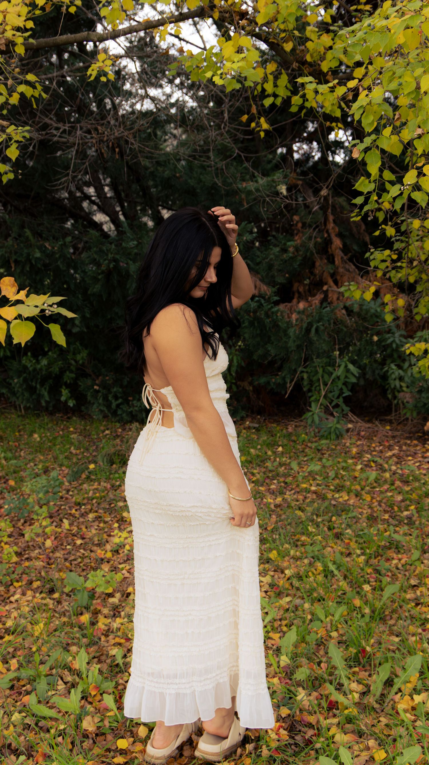 Young woman in cream ruched maxi dress standing among colorful autumn fallen leaves outdoors.