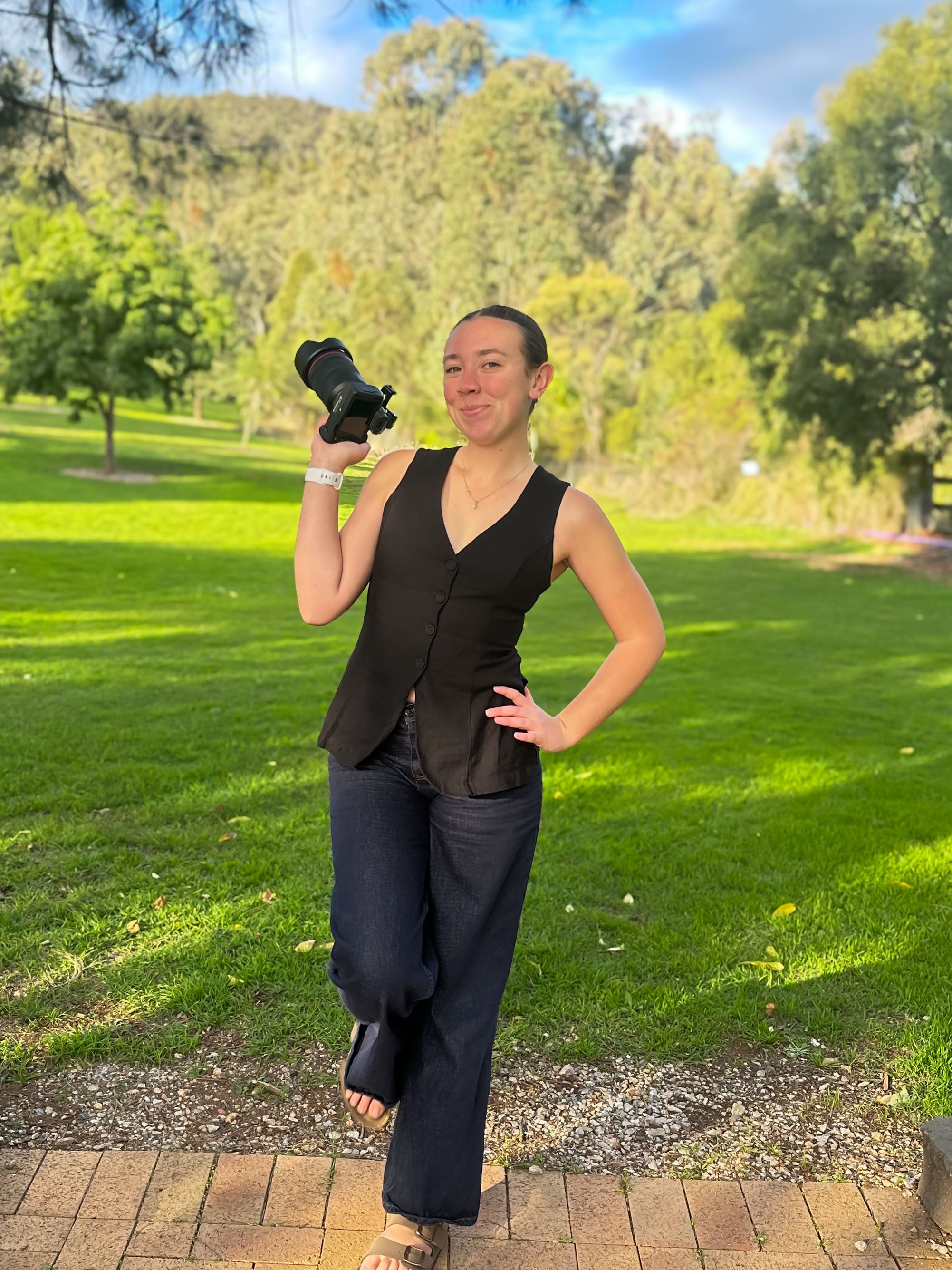 Woman in black vest and pants holds camera in lush green park, smiling confidently on a sunny day.