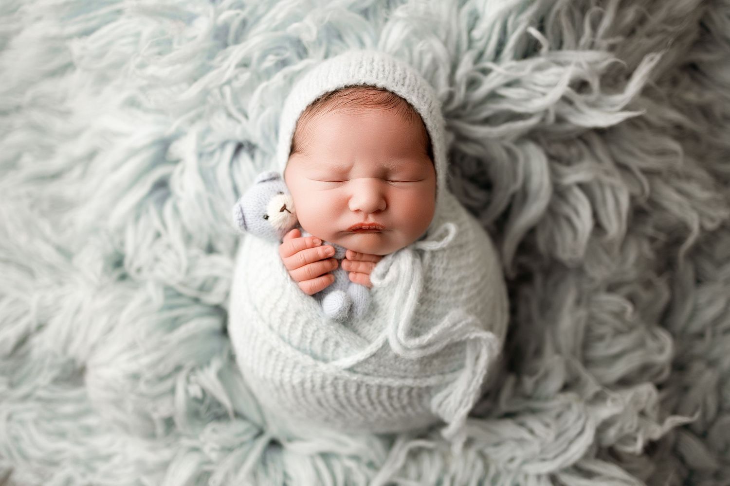Peaceful newborn baby sleeping wrapped in white knit swaddle, wearing bonnet, holding tiny toy on soft blue fur blanket.