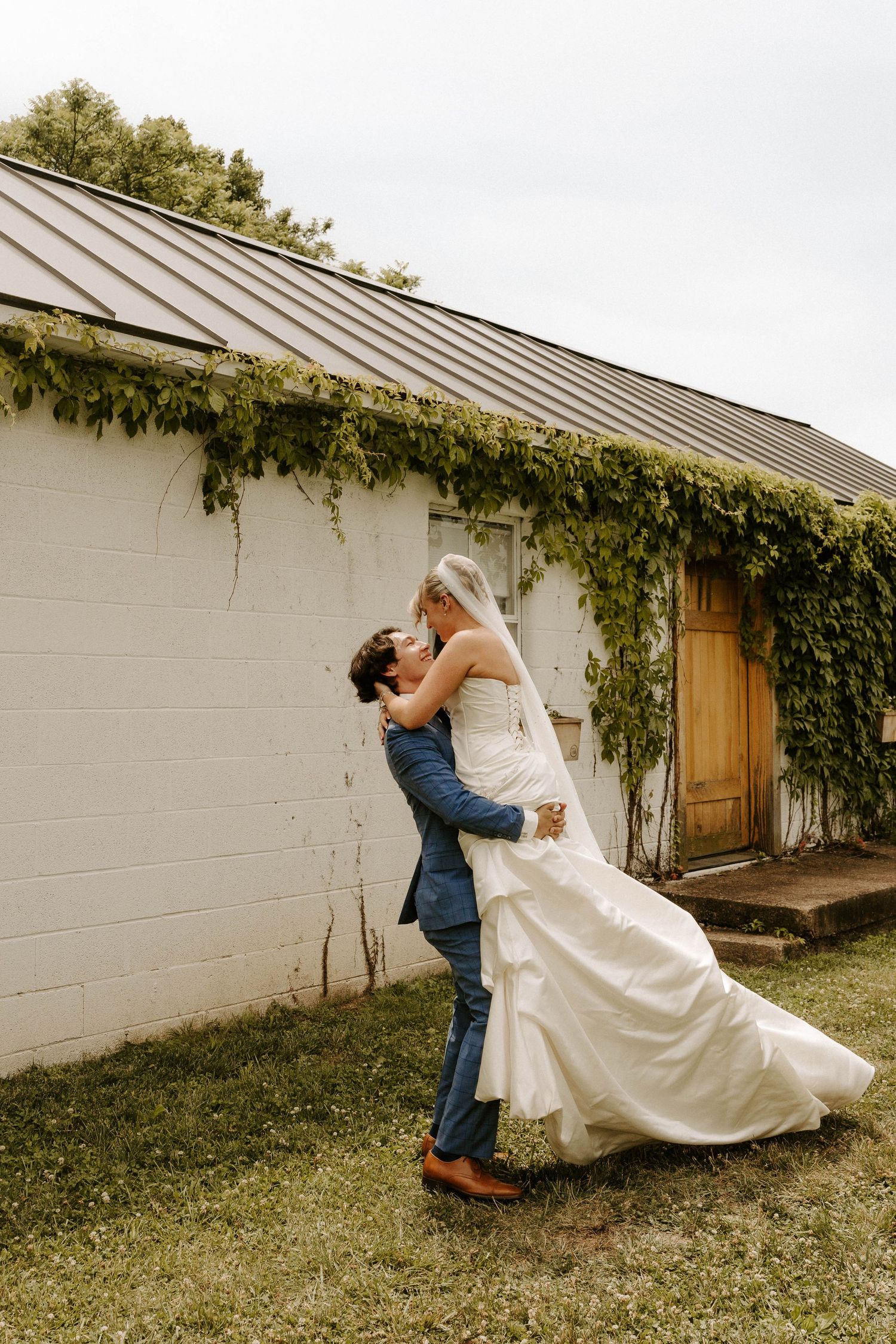 Groom lifts bride in white wedding dress outside rustic white barn covered in ivy, sharing a romantic kiss.