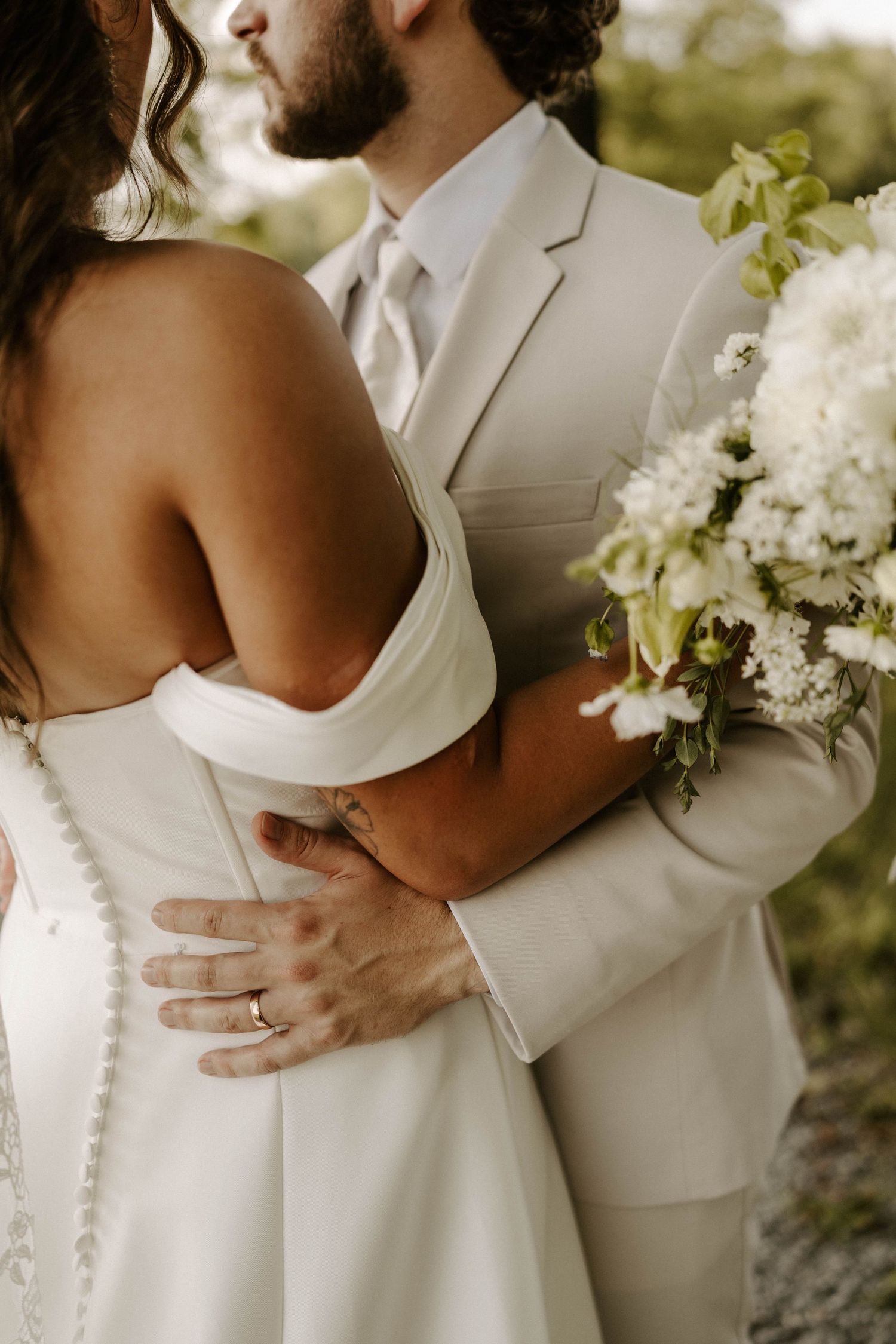 Groom lifts bride in white wedding dress outside rustic white barn covered in ivy, sharing a romantic kiss.