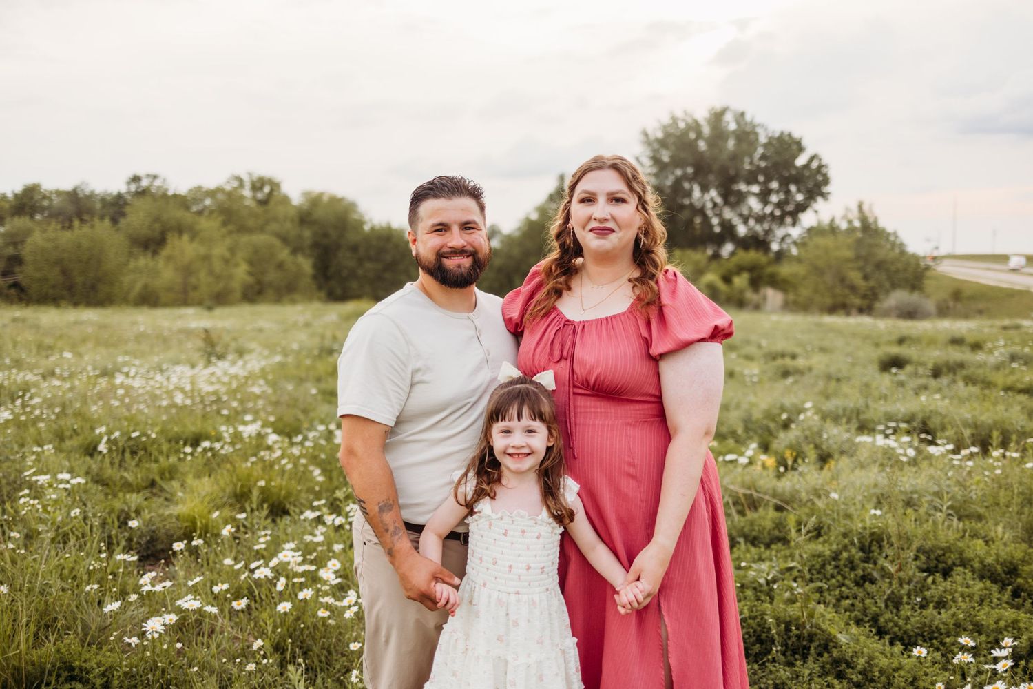 A happy family of three poses in a wildflower field, with a pregnant woman in red, a man, and a young girl.