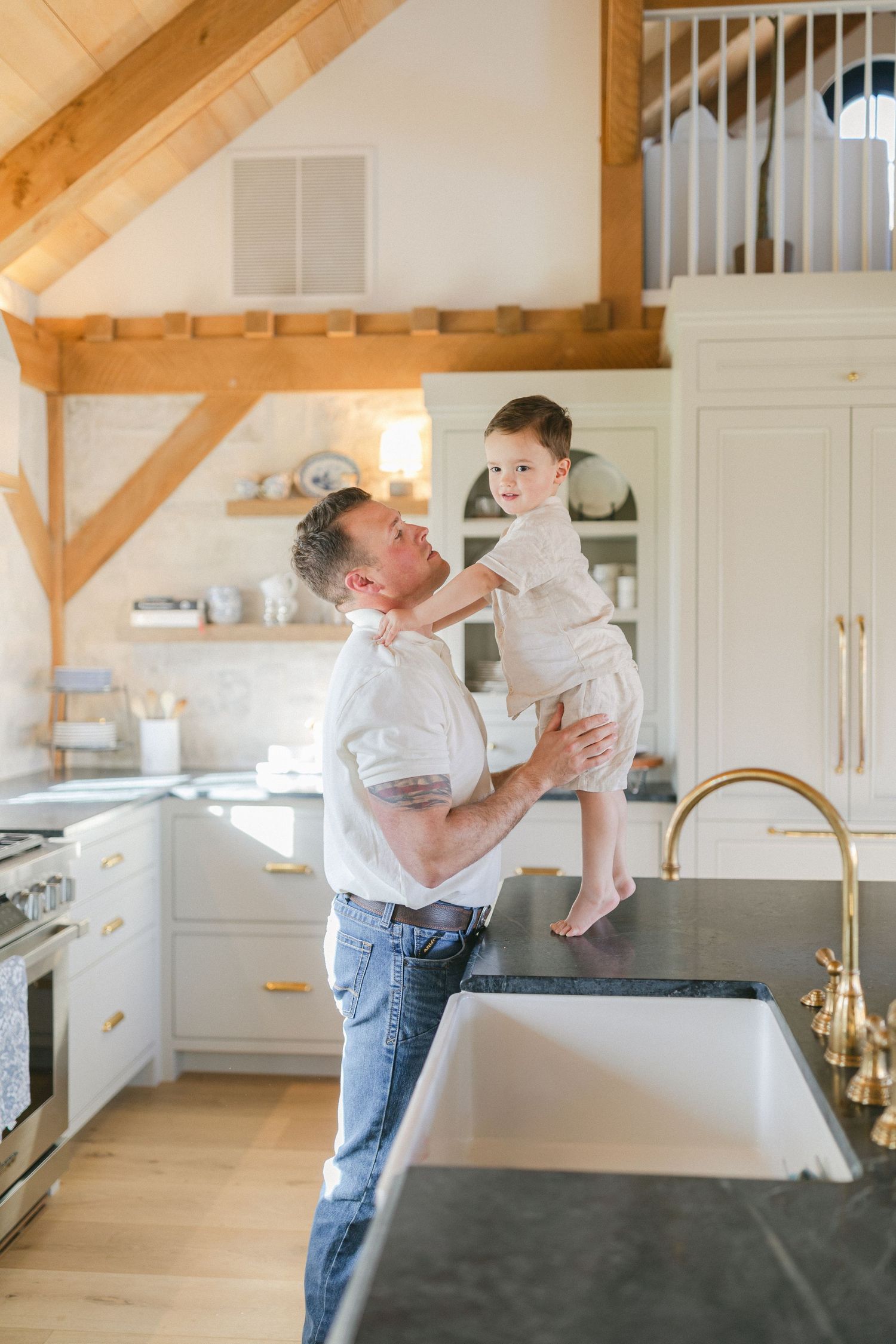 A father lifts his young son in a bright farmhouse kitchen with wood beams, white cabinets, and brass fixtures.