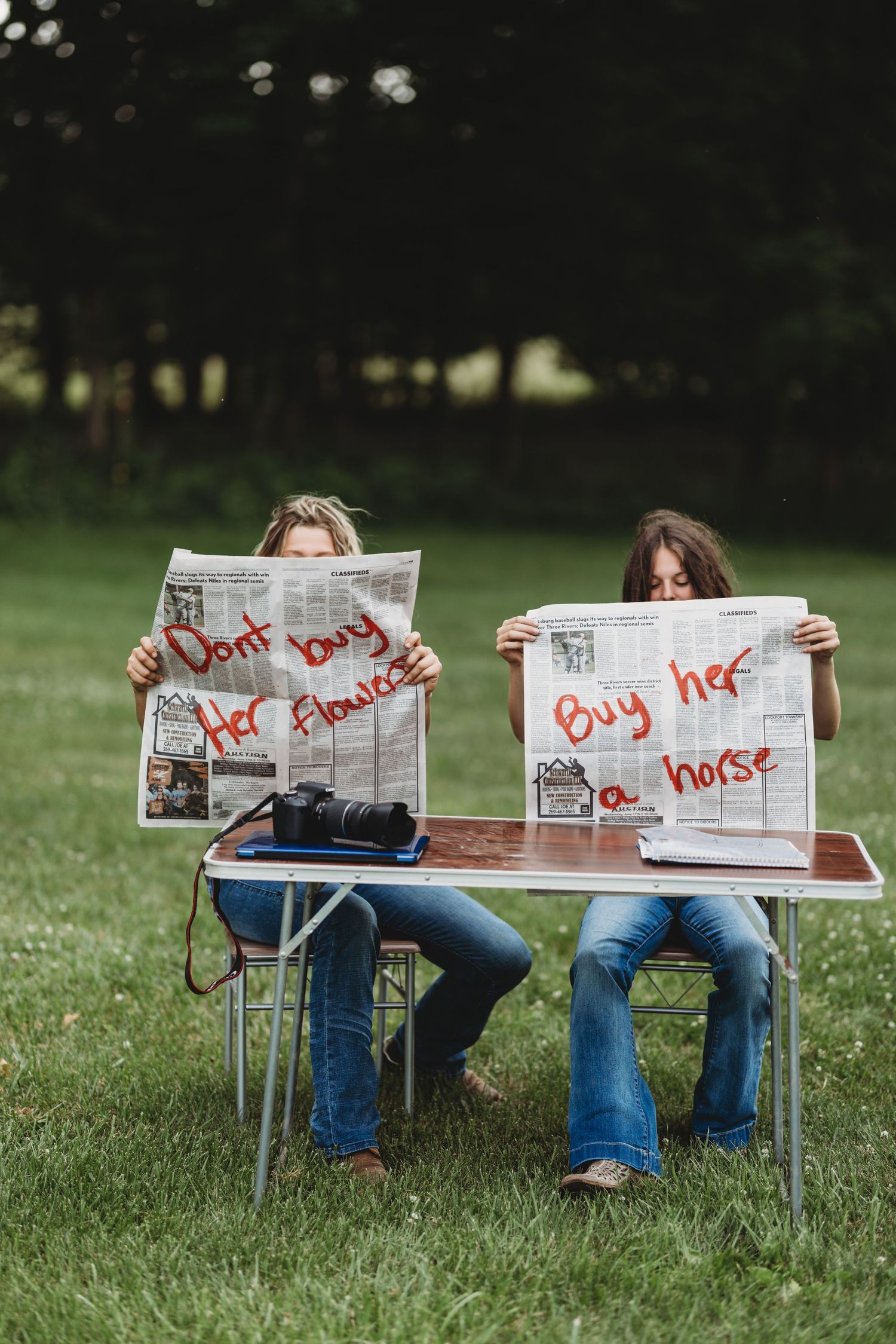 Two women sit outdoors holding newspapers with handwritten messages: "Don't buy her flowers" and "Buy her a horse".