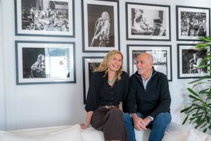 A smiling man and woman sit together on a white sofa surrounded by black and white framed music photos on a white wall.