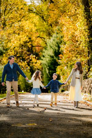 Family of four holding hands walking on a tree-lined path surrounded by vibrant autumn foliage.