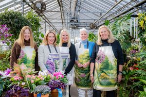Five women wearing garden-themed aprons smile together inside a lush greenhouse surrounded by colorful flowers.