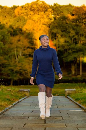 A smiling young Black woman walks along a stone path in a navy sweater dress and cream boots, with autumn trees in the background.