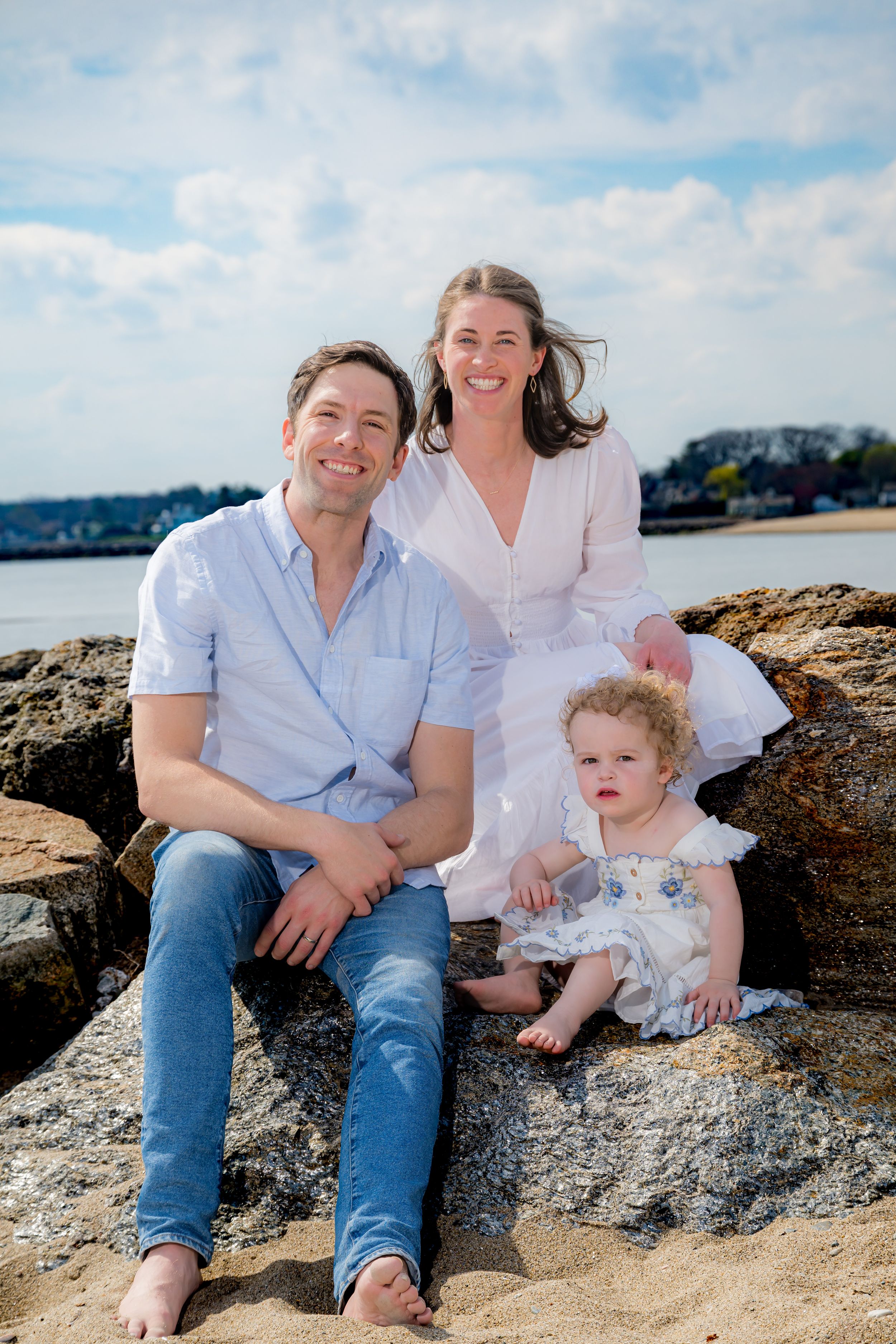 A happy family of three sits on rocky beach, parents smiling with curly-haired toddler, ocean backdrop on sunny day.