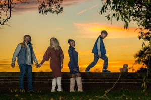 Family of four holding hands at golden sunset on a farm, silhouetted against vibrant orange sky.