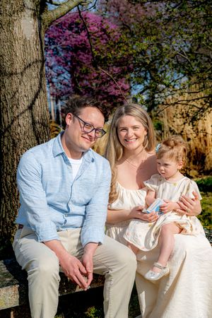 A happy young family with a toddler girl sits outdoors near blooming trees on a sunny spring day.