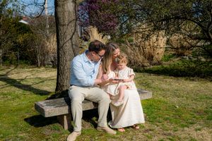 Family of three sitting on a park bench, parents admiring their toddler daughter outdoors in spring.